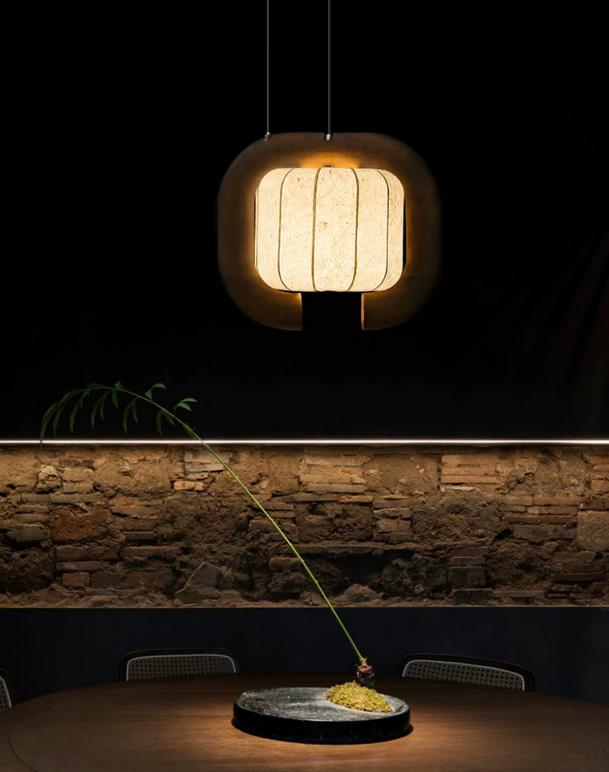 Glowing bright against a dark stone wall, this dark-finished, closed-loop Clérachic pendant lights a table set with a single plate.