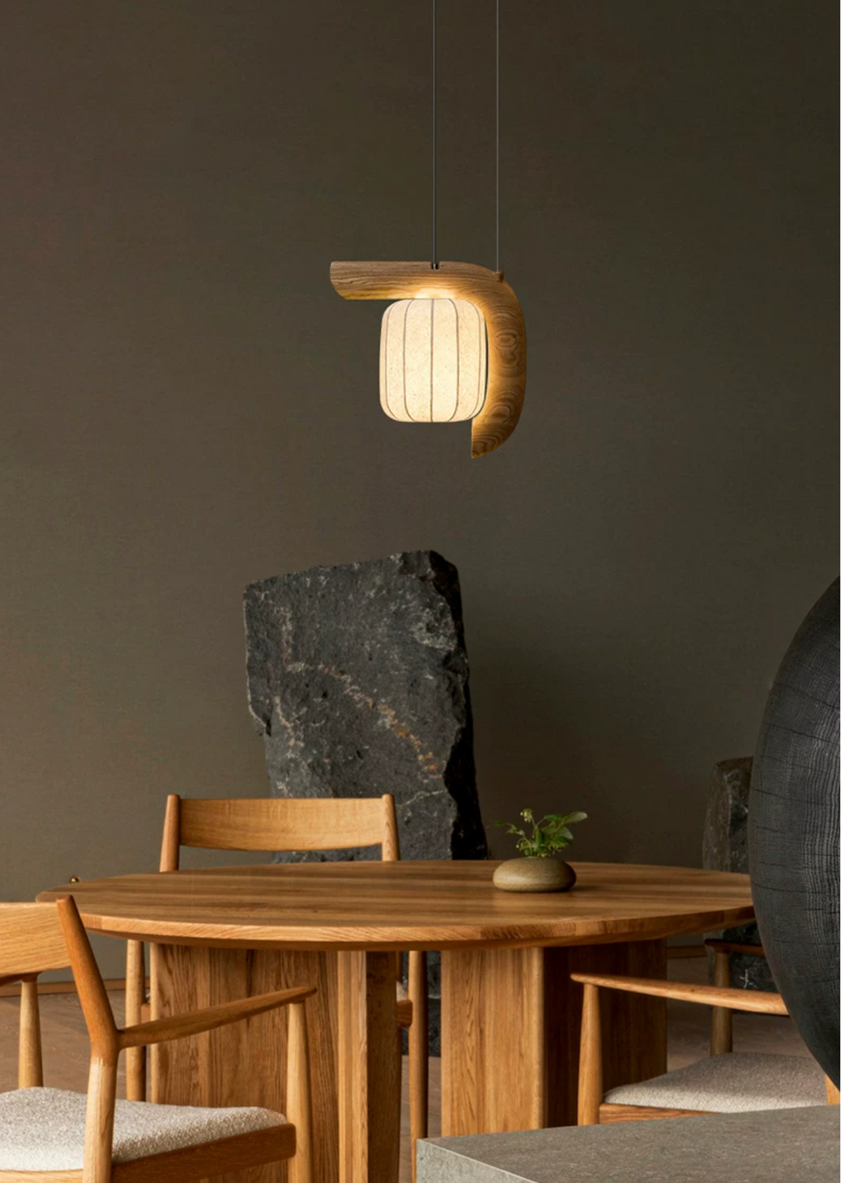 This dark wood, bent-style Clérachic pendant hangs above a round wood dining table, a stone sculpture in the background.