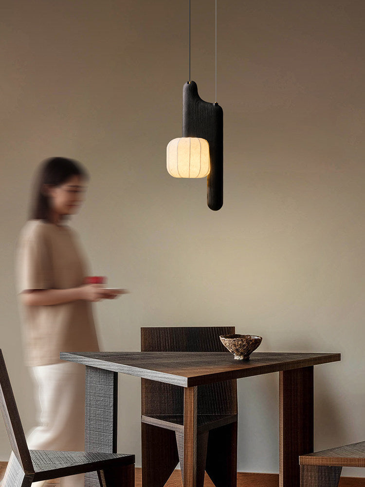 This black, vertical-style Clérachic pendant glows above a wood table as a figure walks past in soft focus.
