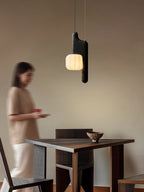 This black, vertical-style Clérachic pendant glows above a wood table as a figure walks past in soft focus.