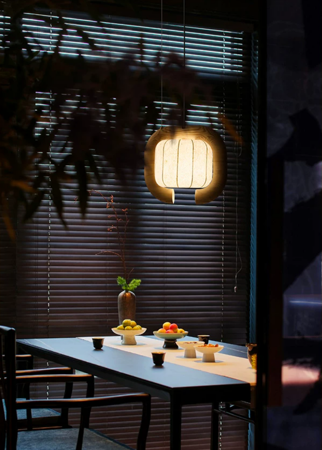 In front of louvred blinds, this dark-finished, closed-loop Clérachic pendant glows above a table set with bowls of fruit.