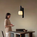 This black, vertical-style Clérachic pendant glows in a plain room as a figure walks past in the background.