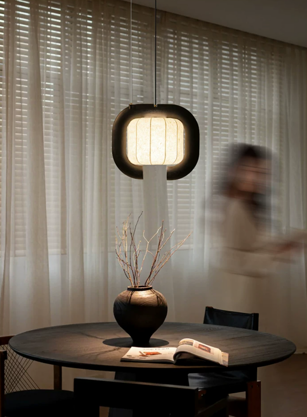 In a dimly lit room, this dark-finished, closed-loop Clérachic pendant glows above a round table set with a vase.