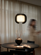 In a dimly lit room, this dark-finished, closed-loop Clérachic pendant glows above a round table set with a vase.