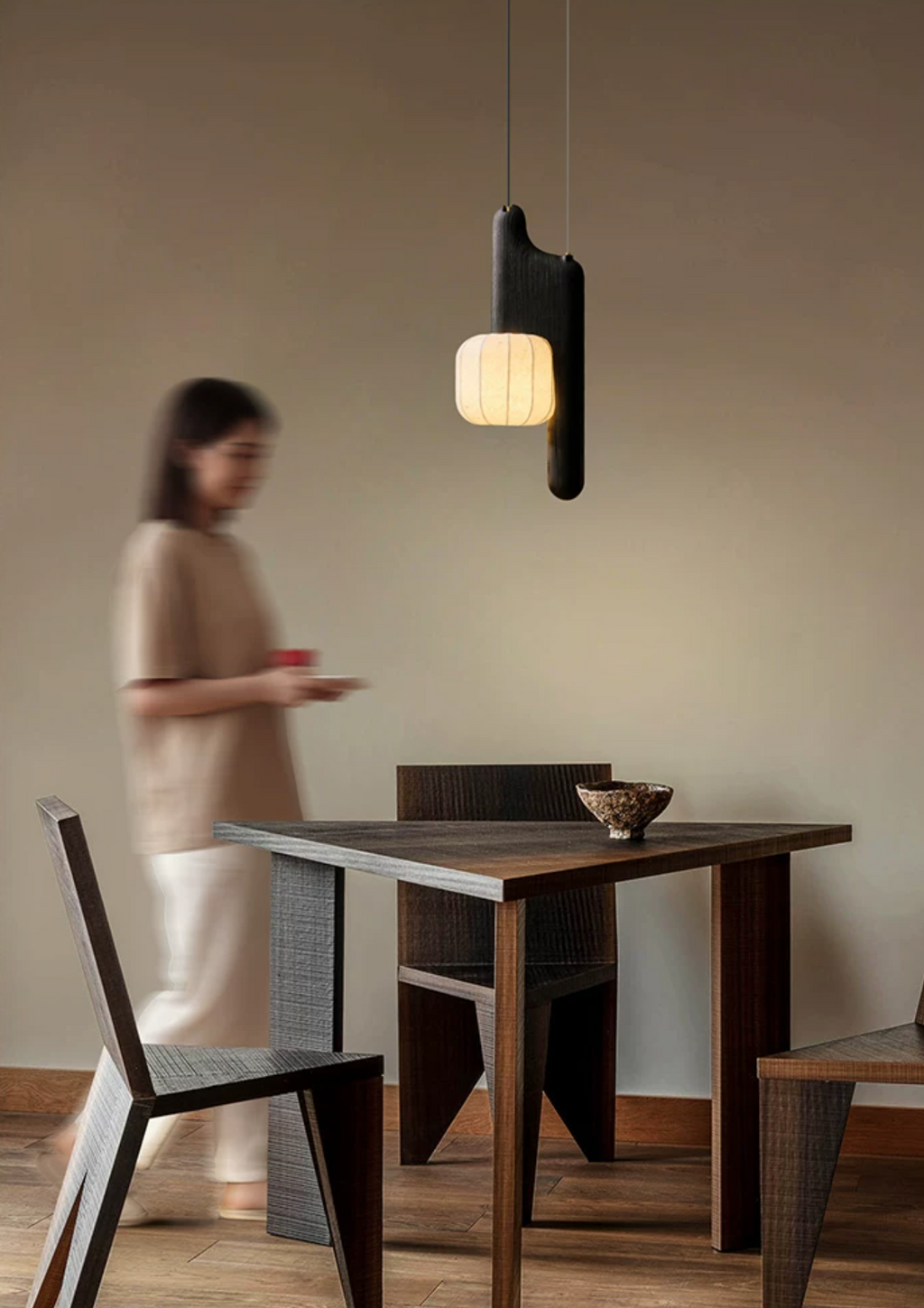 This black, vertical-style Clérachic pendant glows above a small square table as a figure walks past nearby.
