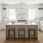Grey cabinetry and wood stools sit beneath this Clérachic pendant, glowing warm above a kitchen island near a window.