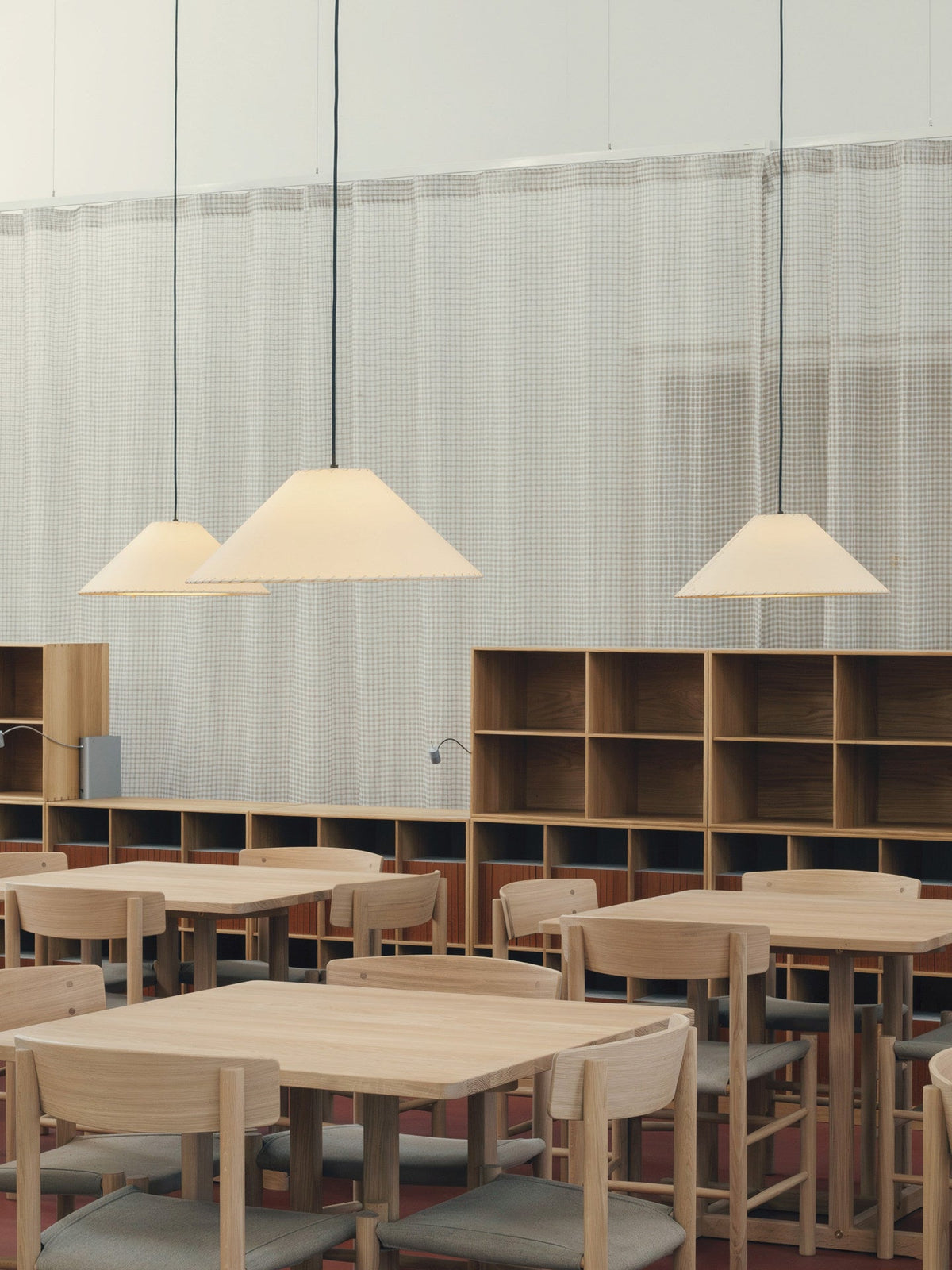Three white Clérachic cone pendants light a row of pale wooden café tables against a sheer curtain.