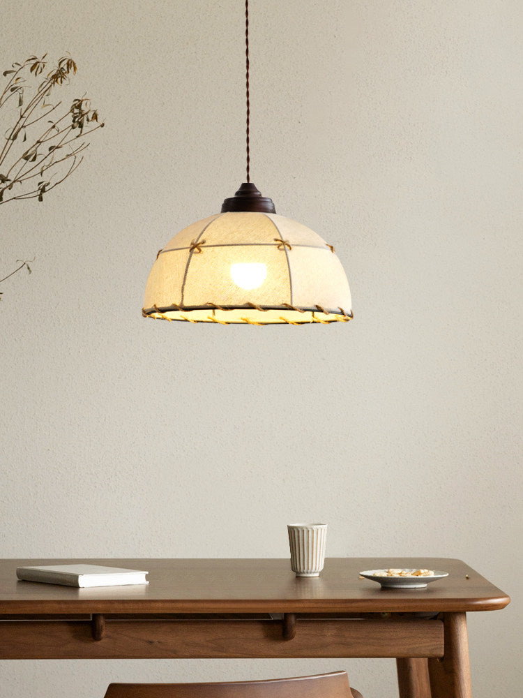 This Clérachic pendant's quilted beige shade glows above a wood table set with a book and cup, near a dried branch.