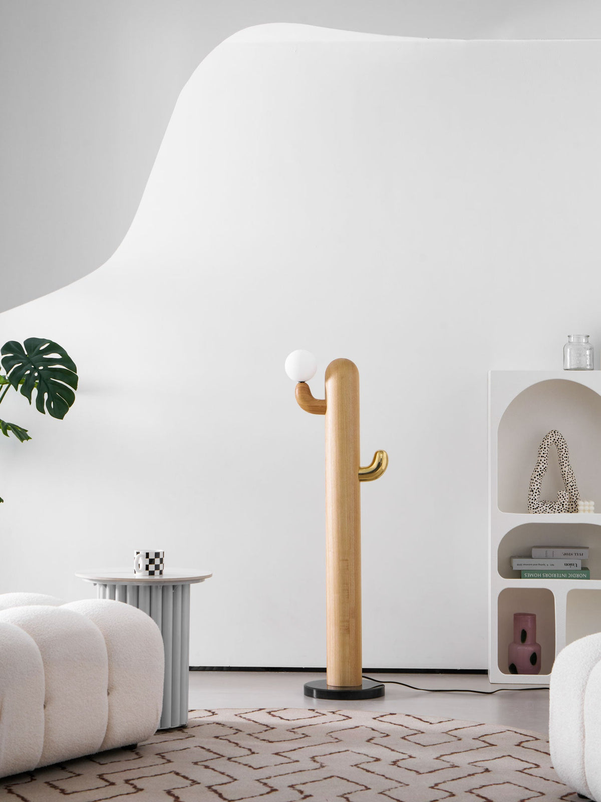 Clérachic wood floor lamp beside a full leafy plant, its brass-hooked glass globe unlit against the arched bookshelf backdrop.