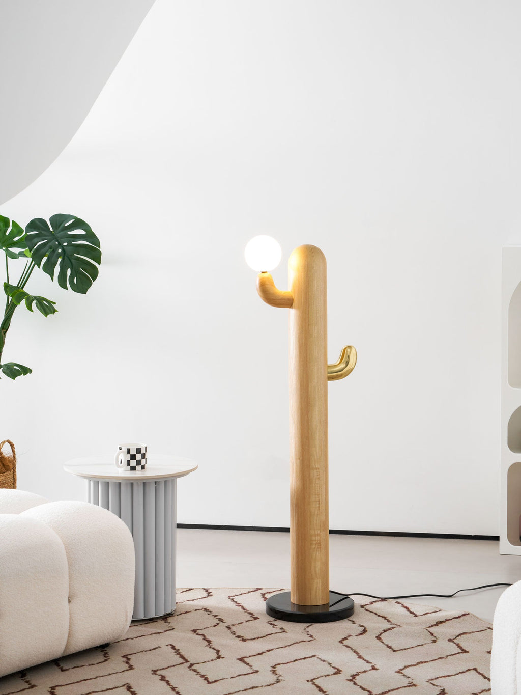 Clérachic wood floor lamp lit warm beside a tall leafy plant, the arched bookshelf and its trinkets filling the room behind.