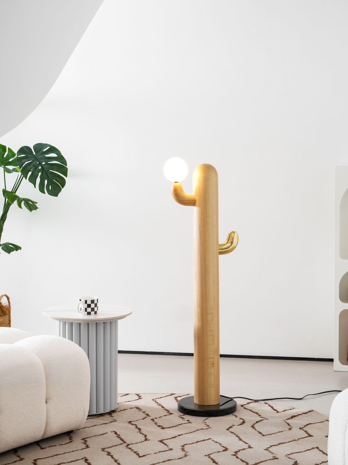 Clérachic wood floor lamp lit warm beside a tall leafy plant, the arched bookshelf and its trinkets filling the room behind.