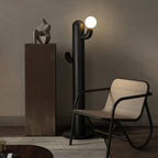 Black-finished Clérachic floor lamp lit beside a small sculpture on a bronze pedestal and a black cane armchair.