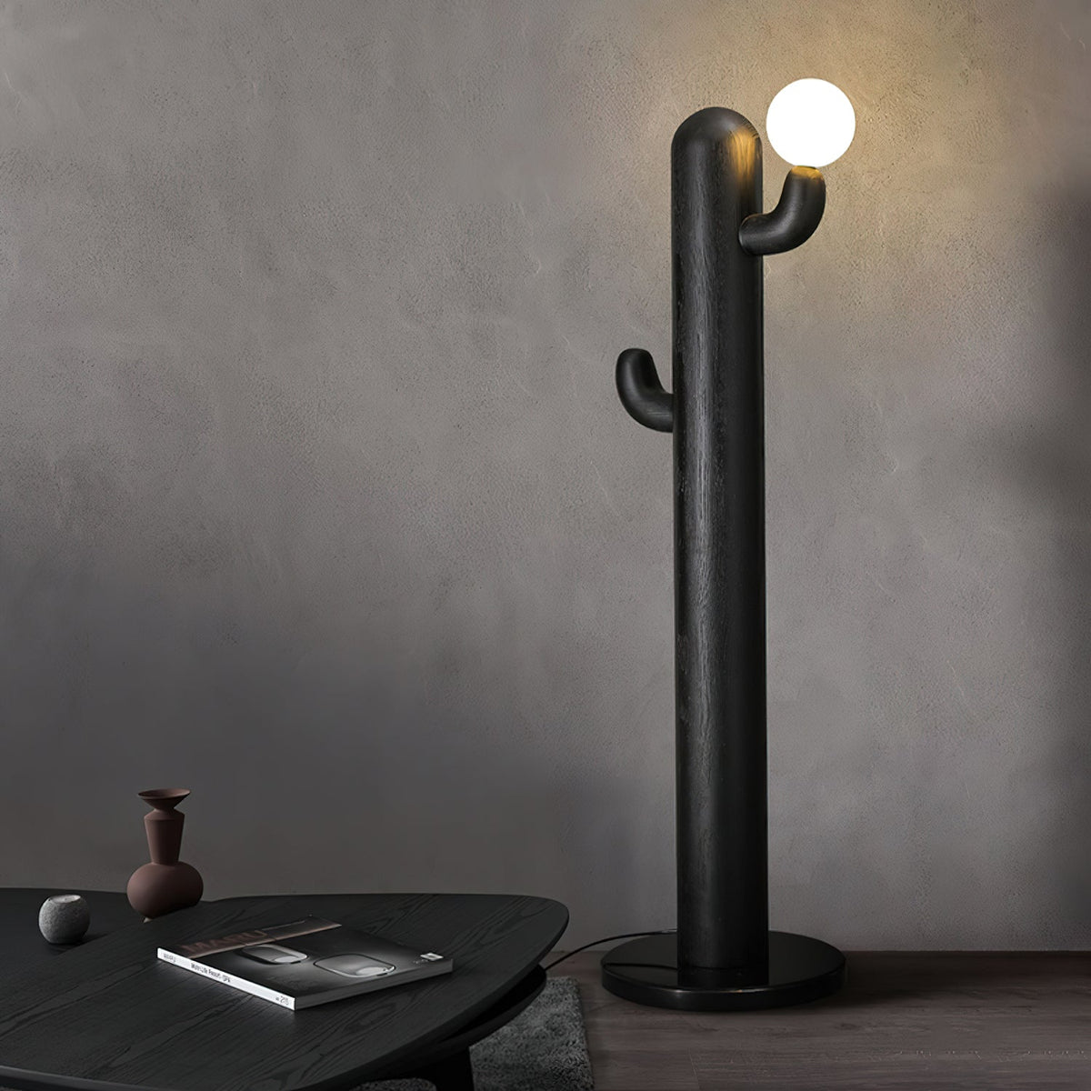 Black Clérachic floor lamp glows against a grey wall beside a dark round coffee table stacked with books.