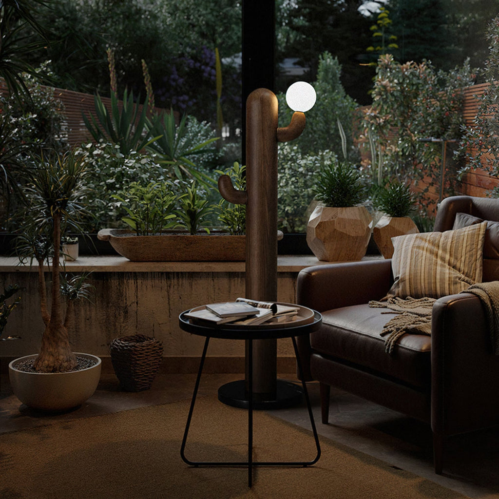 Dark wood Clérachic floor lamp glows on an evening patio beside potted plants and a round table with books.