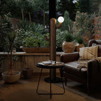 Dark wood Clérachic floor lamp glows on an evening patio beside potted plants and a round table with books.