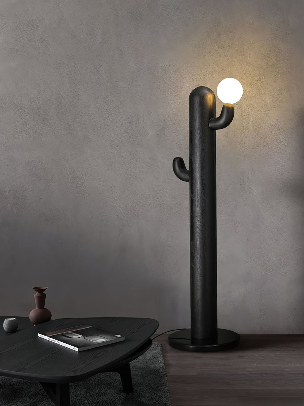 Black Clérachic floor lamp glows against a plain grey wall beside a round black table holding a book and small vase.