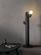 Black Clérachic floor lamp glows against a plain grey wall beside a round black table holding a book and small vase.