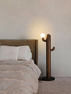Dark wood Clérachic floor lamp lit beside a tan headboard and blush pink bedding in a warm-toned bedroom.