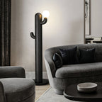 Black Clérachic floor lamp glows beside a curved grey sofa with dark pillows and a round table holding a ceramic bowl.