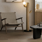 Light wood Clérachic floor lamp glows beside a woven wood armchair, a round black table and a shelf of books.