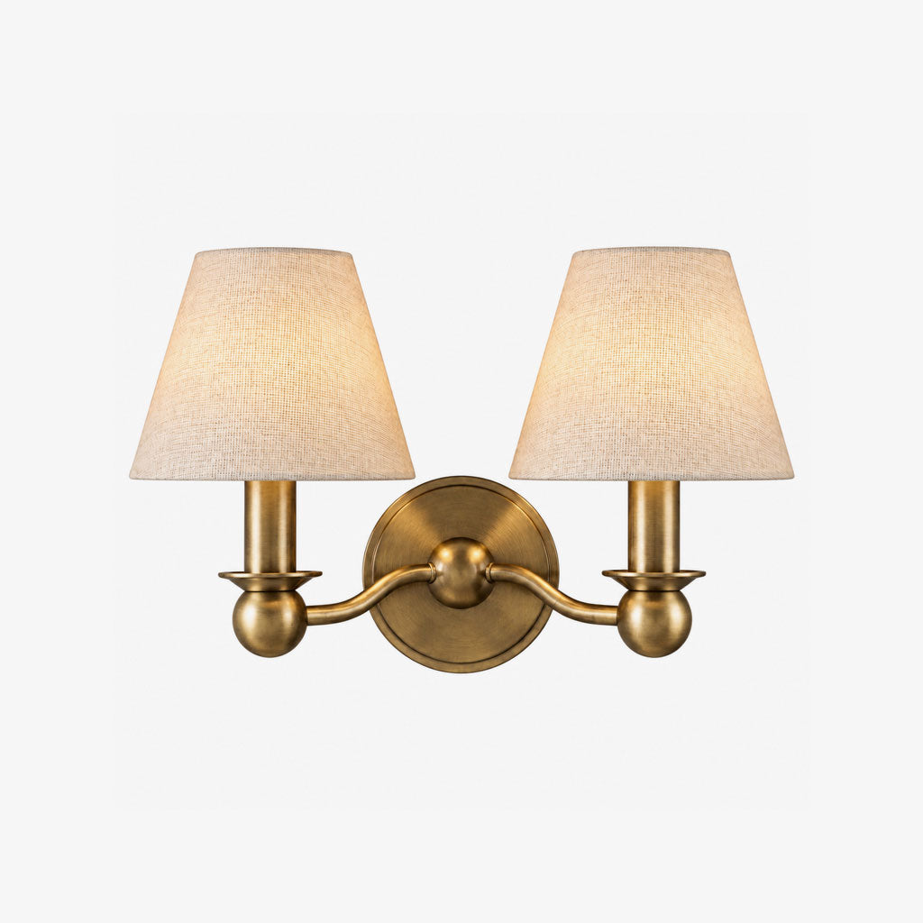 A brushed brass Clérachic wall sconce with twin fabric drum shades, photographed unlit on a plain backdrop.