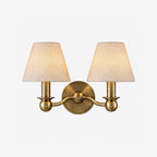 A brushed brass Clérachic wall sconce with twin fabric drum shades, photographed unlit on a plain backdrop.