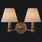 This Clérachic wall sconce shows its double-arm fabric drum shades in brushed brass, unlit against a dark backdrop.