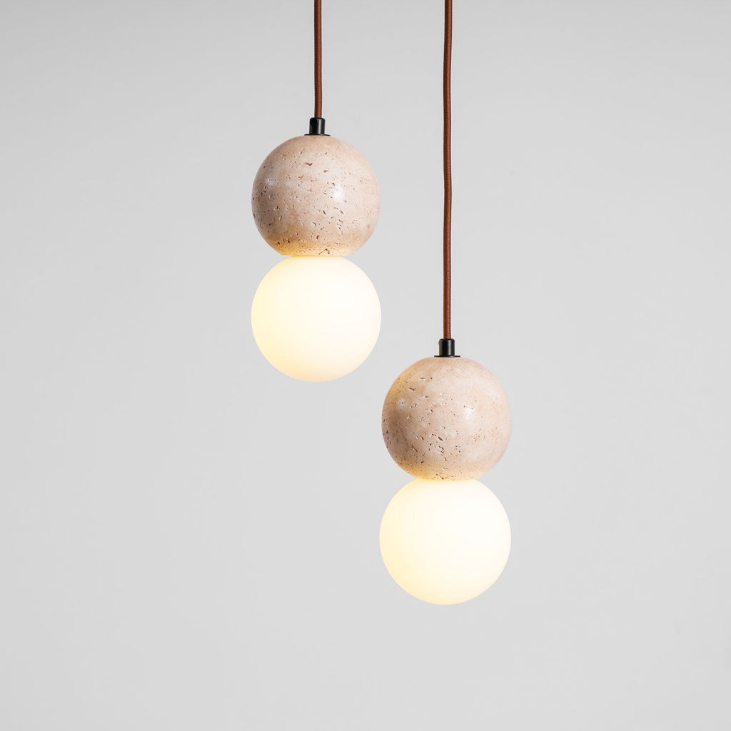 Two lit Clérachic pendants hang side by side, each a travertine sphere paired with a glowing opal glass globe.