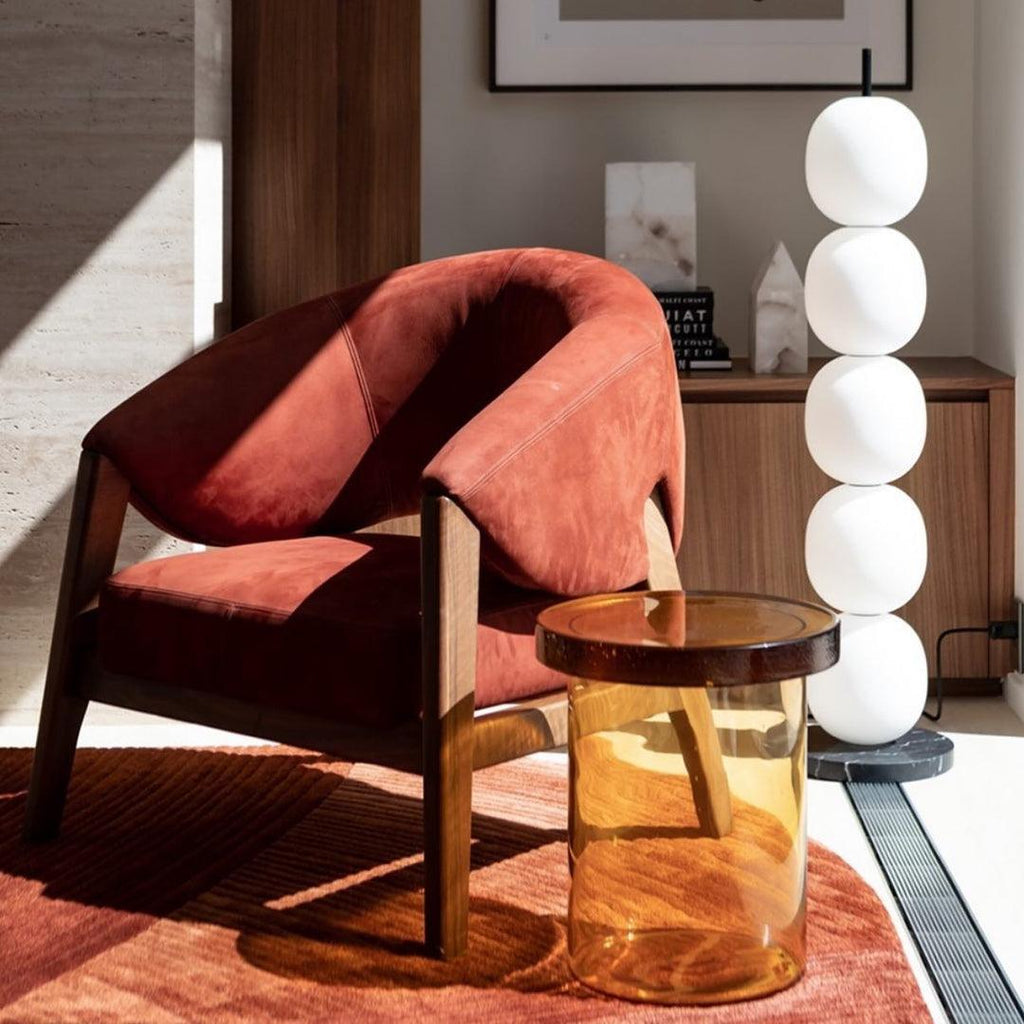 The lamp unlit beside a rust suede armchair and an amber glass side table in low afternoon sun.