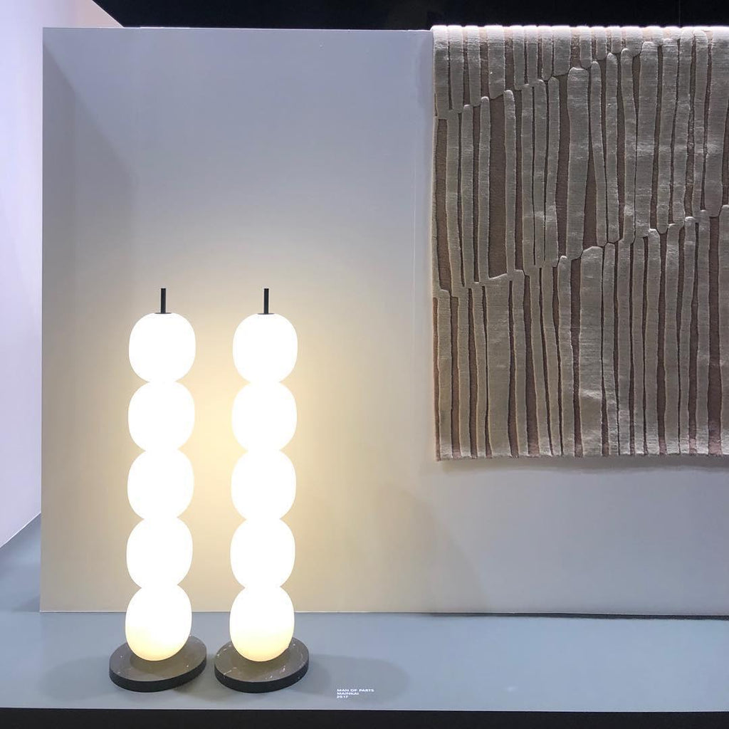 Two lamps lit side by side on a pale plinth, next to a striped rug hung on the wall.