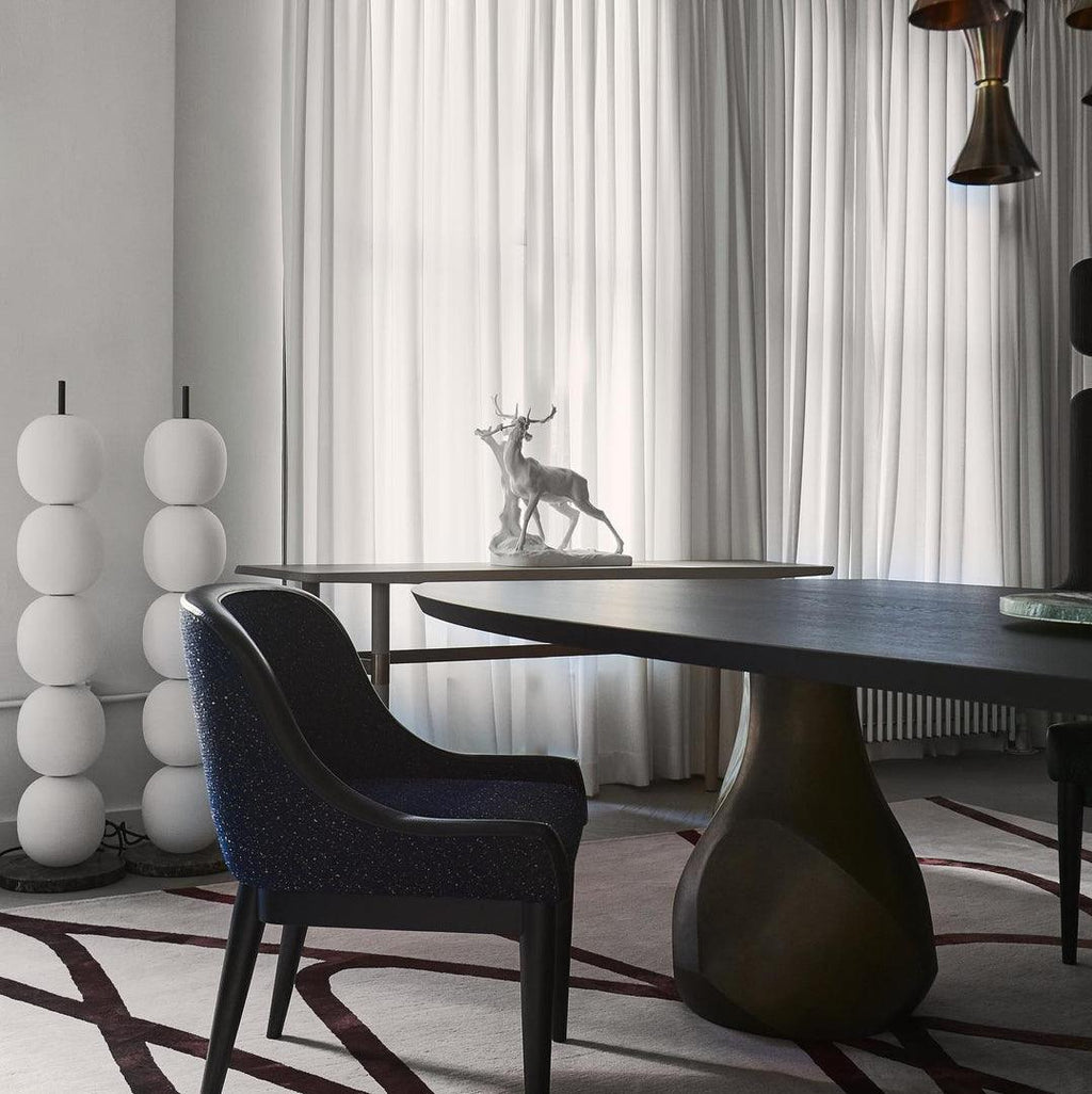 Two unlit lamps standing together in a corner by white curtains, behind a dark dining table.