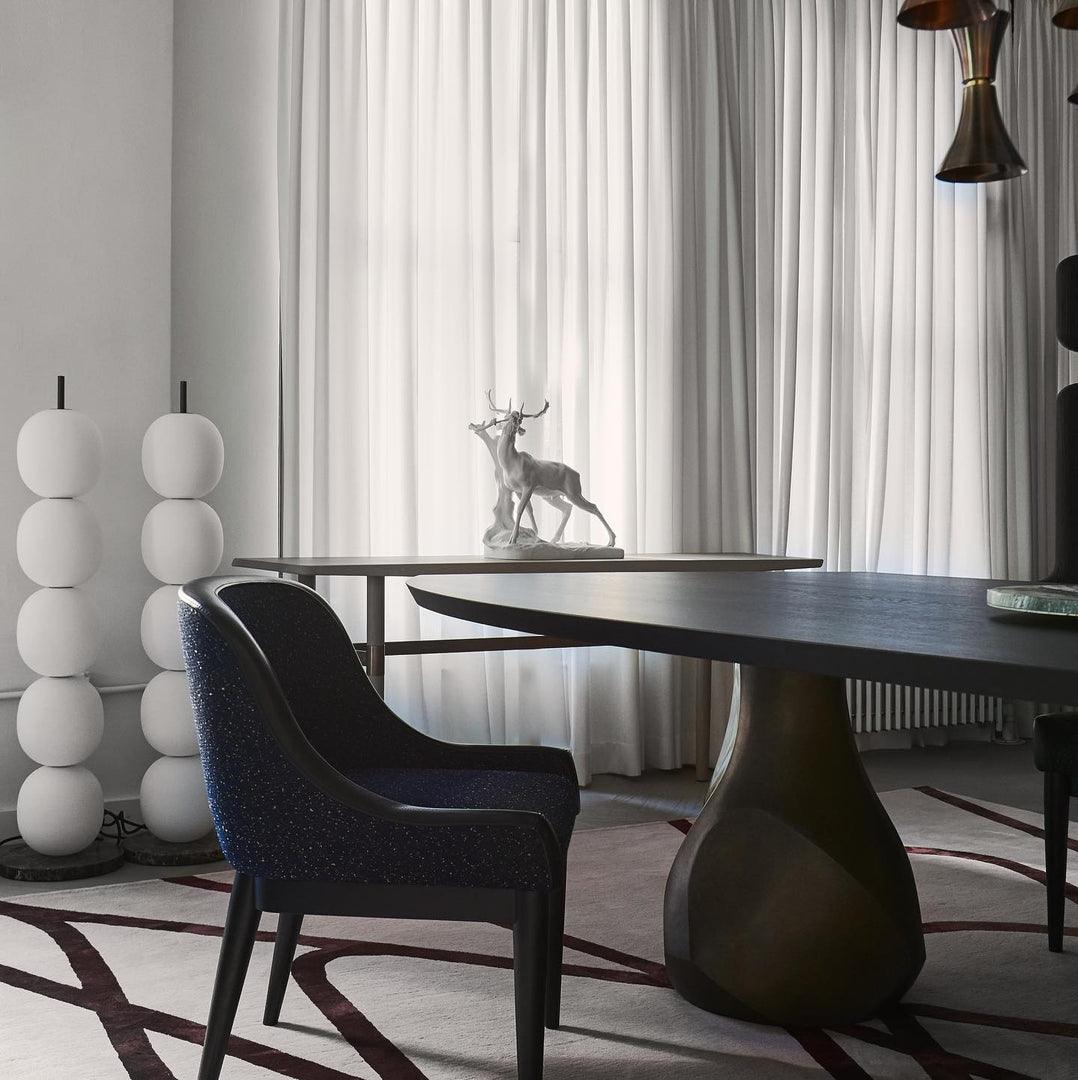 Two unlit lamps standing together in a corner by white curtains, behind a dark dining table.