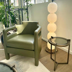 Lit lamp behind an olive leather armchair and a bronze and glass side table.