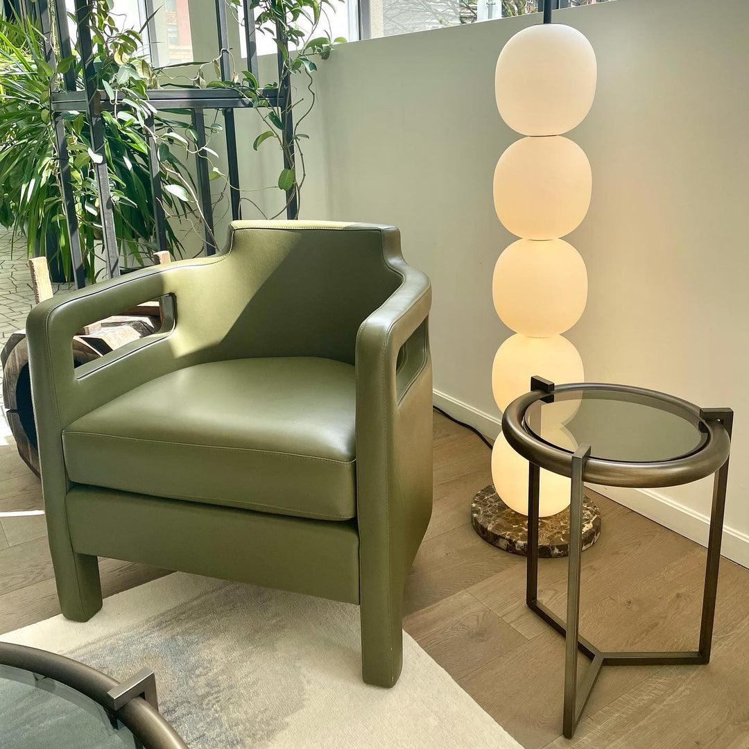 Lit lamp behind an olive leather armchair and a bronze and glass side table.