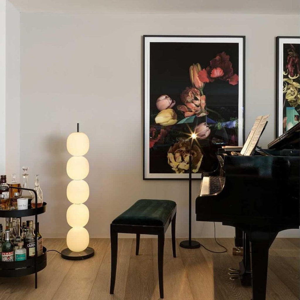 Lit lamp beside a grand piano and a drinks trolley, under a large framed floral print.
