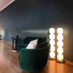 Two lit lamps side by side against a dark teal wall, beside a green velvet sofa.