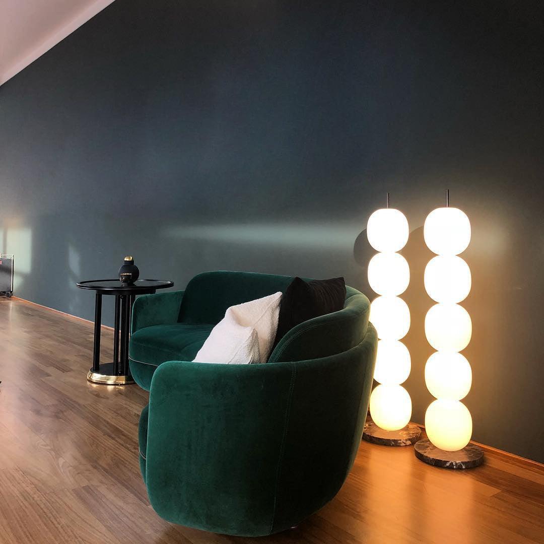 Two lit lamps side by side against a dark teal wall, beside a green velvet sofa.