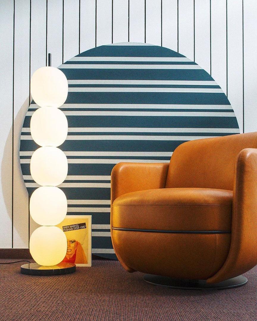 Lit lamp next to a tan leather swivel chair, a round striped backdrop and a propped art book behind.