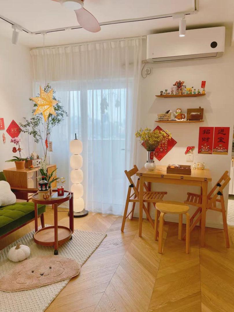 The lamp standing at a sheer curtain beside a green sofa, with a dining table opposite.