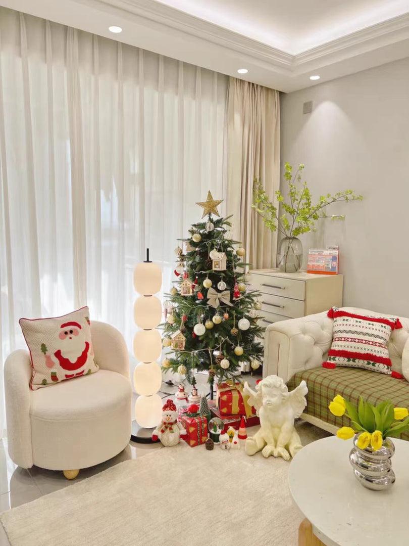 Lit lamp beside a boucle armchair and a decorated tree in a pale living room.