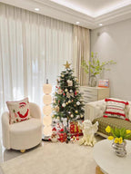 Lit lamp beside a boucle armchair and a decorated tree in a pale living room.
