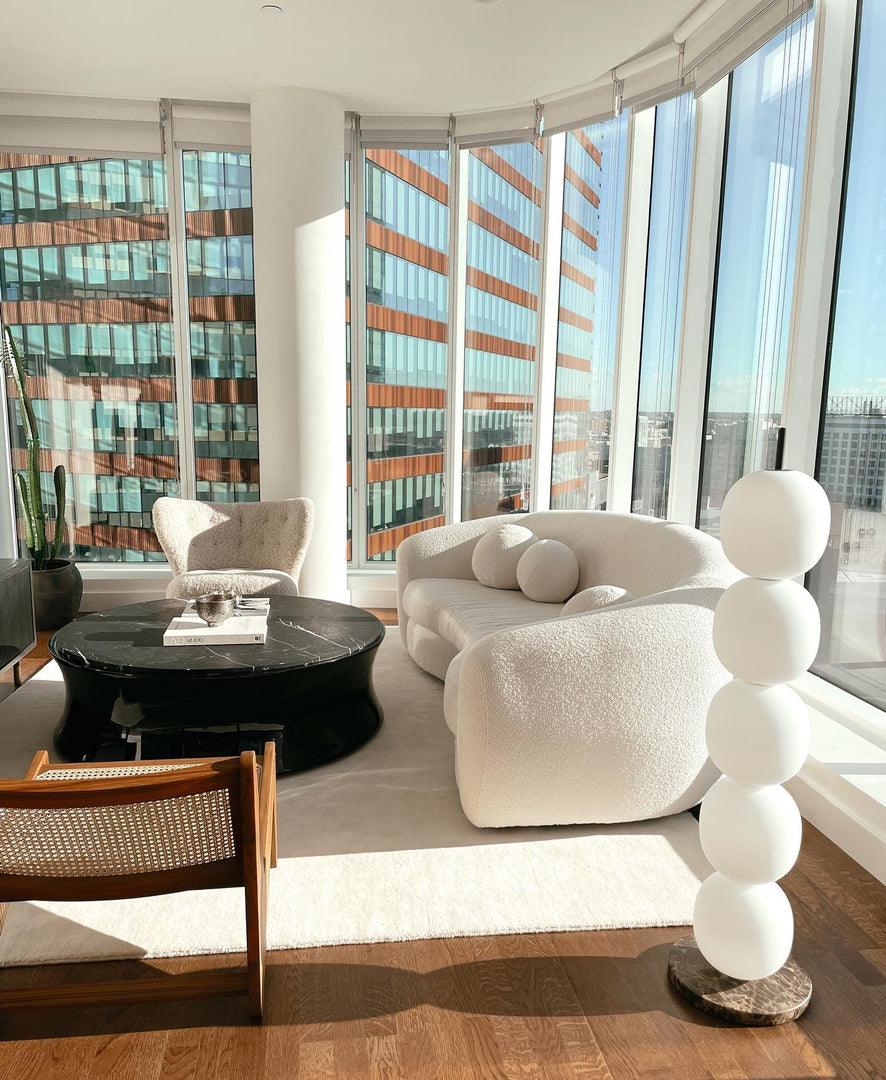 Unlit lamp on a brown marble base by a curved window wall, beside a white boucle sofa.