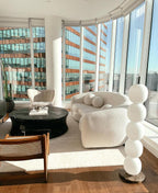 Unlit lamp on a brown marble base by a curved window wall, beside a white boucle sofa.