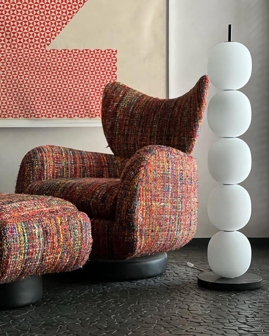 Unlit lamp next to a multicoloured tweed armchair and footstool, on a dark stone floor.