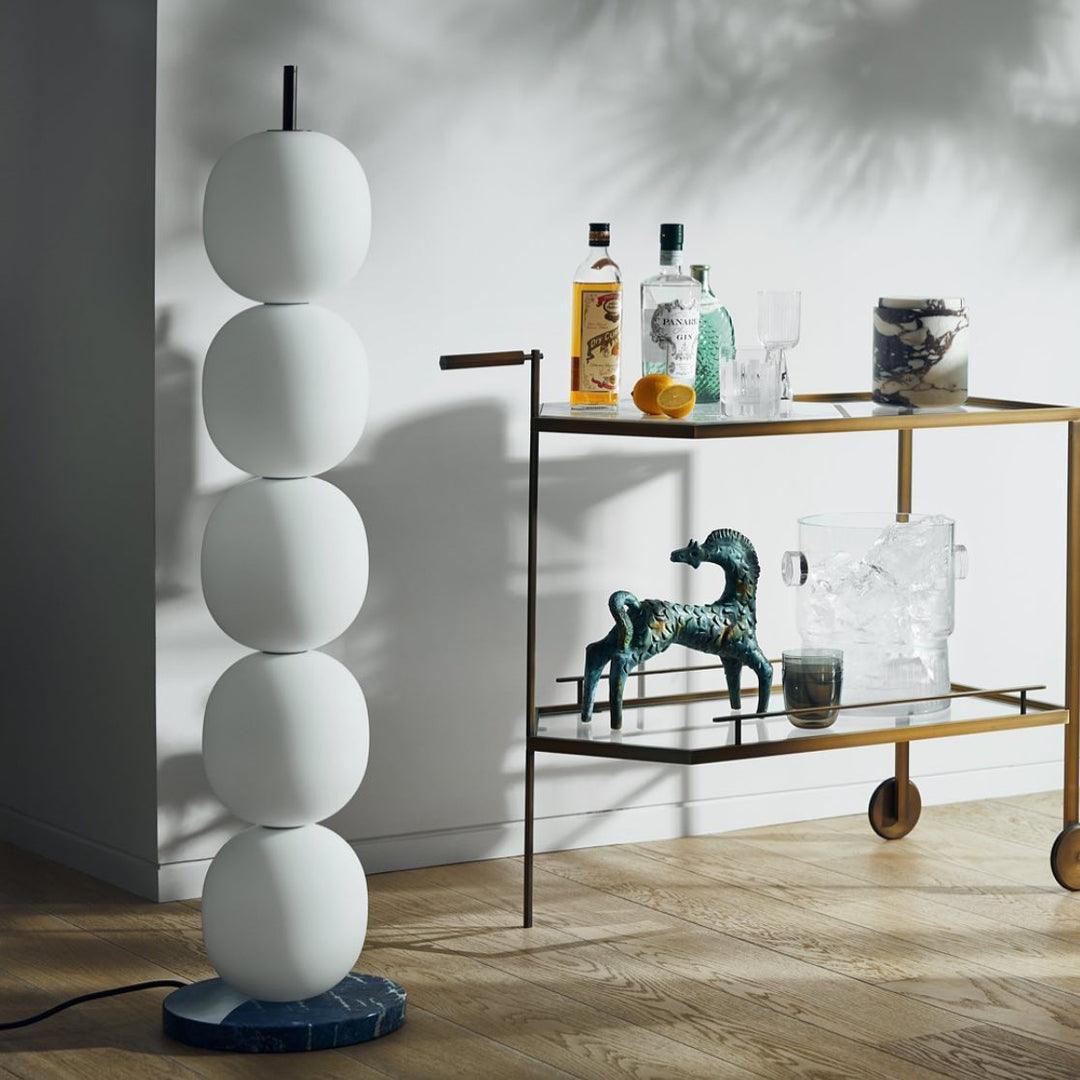 Unlit lamp on a green veined marble base beside a brass drinks trolley, leaf shadows on the wall.
