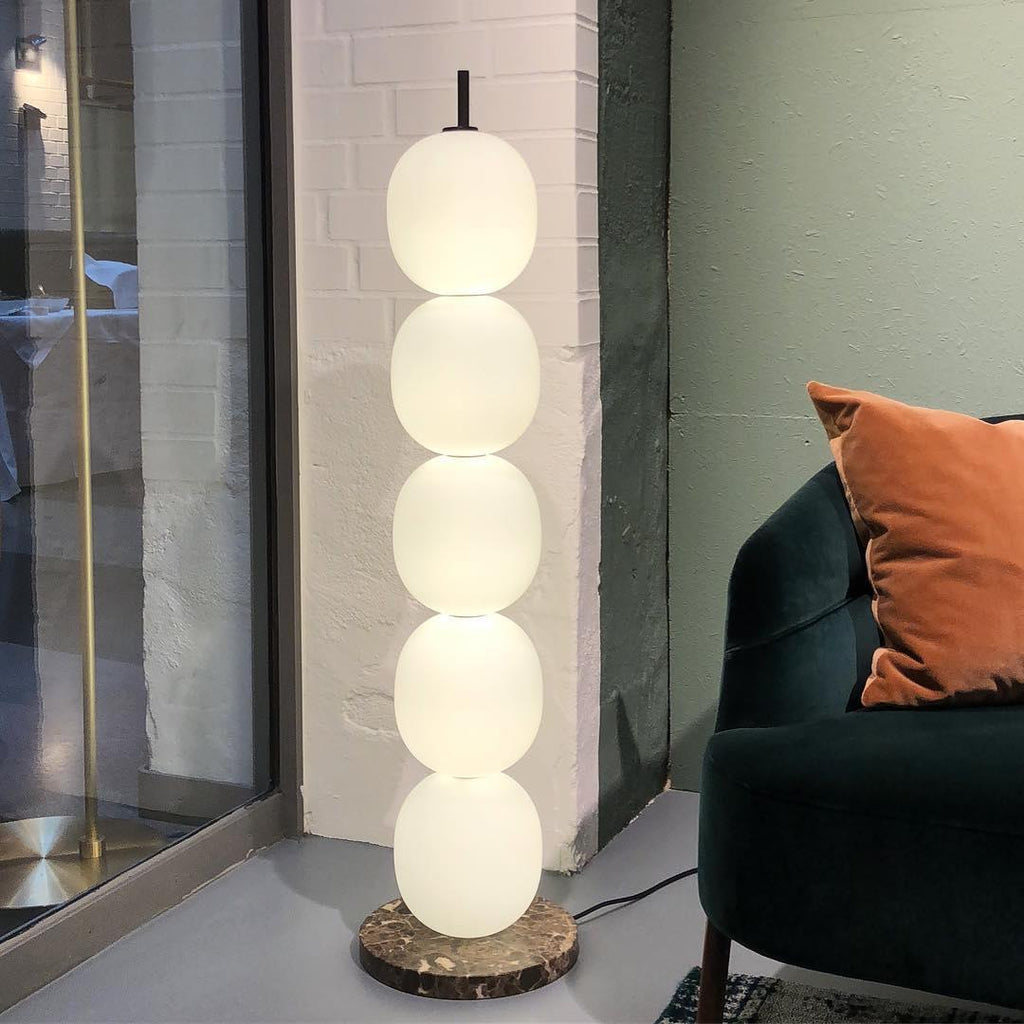 Lit lamp on a red veined marble base against white brick, next to a green velvet chair.