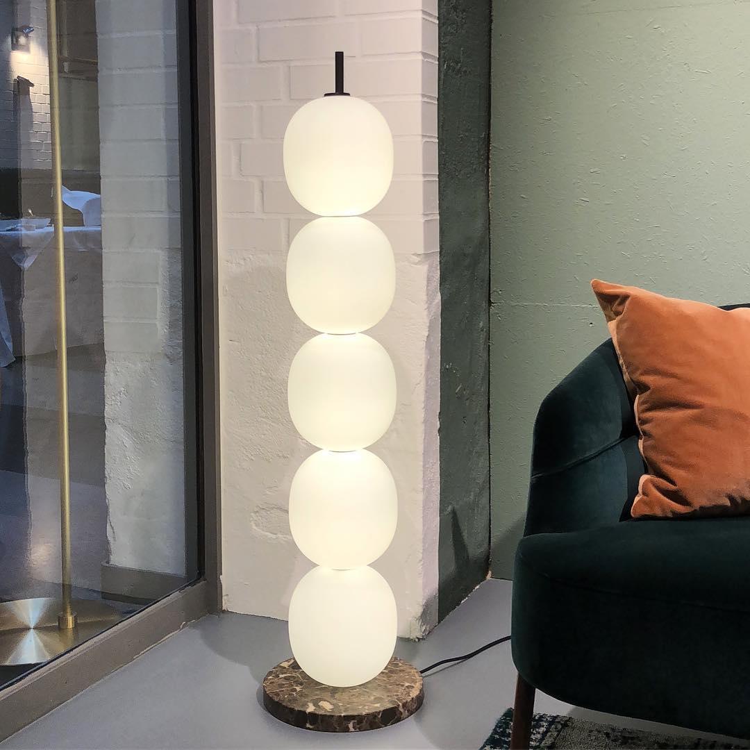 Lit lamp on a red veined marble base against white brick, next to a green velvet chair.