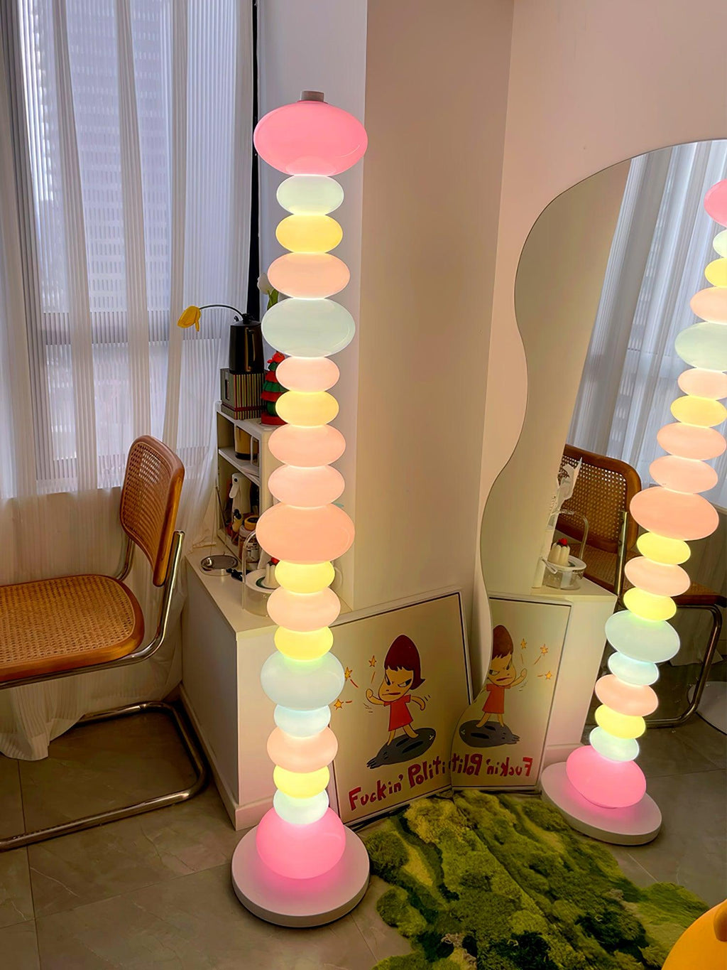 Beside a round mirror and a rattan chair, two Clérachic floor lamps glow pink and multicolour against a patterned wall.