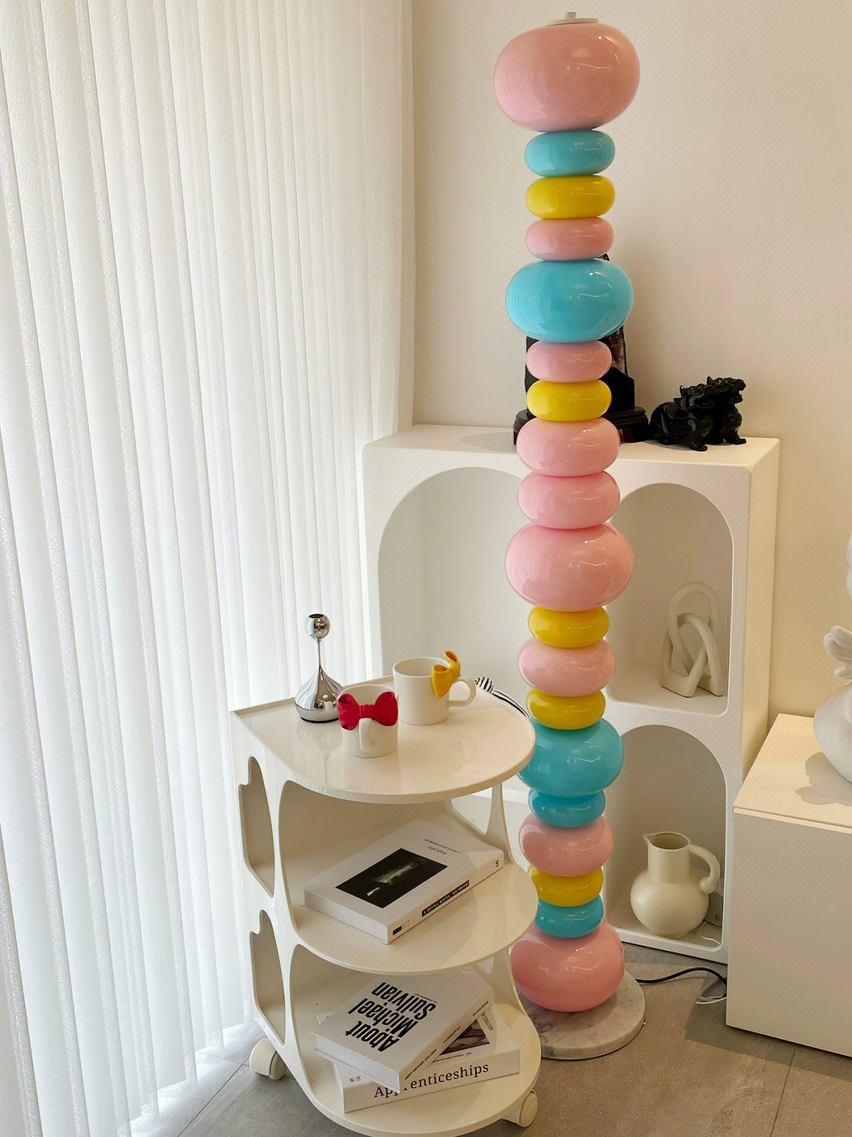 Clérachic multicolour floor lamp lit beside a white shelving unit stacked with books and mugs.