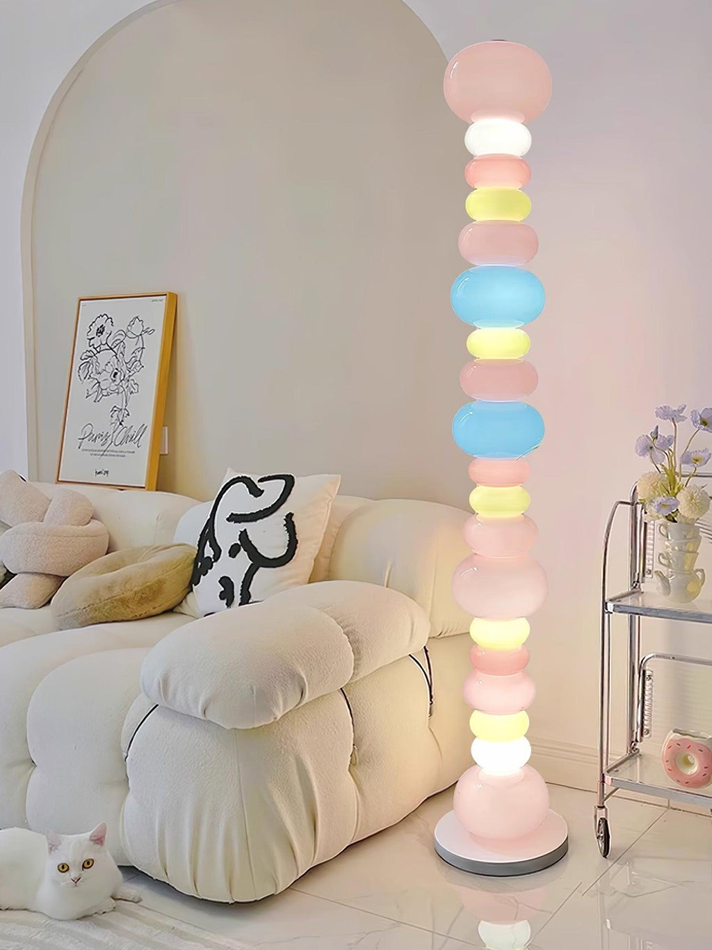 A white cat rests near the base of this Clérachic floor lamp, its lit globes rising beside a cream armchair.